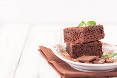 Çikolatalı kek kare parçalar halinde beyaz plaka üzerine nane yaprakları ve beyaz vintage ahşap zemin üzerine kakao tozu ile dekore edilmiştir. Amerikan geleneksel lezzetli tatlı. Fotoğraf kadar kapatın