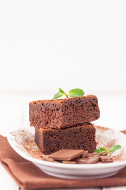 Çikolatalı kek kare parçalar halinde beyaz plaka üzerine nane yaprakları ve beyaz vintage ahşap zemin üzerine kakao tozu ile dekore edilmiştir. Amerikan geleneksel lezzetli tatlı. Fotoğraf kadar kapatın