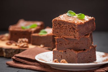 Çikolatalı kek kare parçalar halinde ceviz, beyaz plaka üzerine nane yaprakları ve siyah arka plan üzerine kakao ile dekore edilmiştir. Lezzetli tatlı. Karanlık ruh. Fotoğraf kadar kapatın. Seçici odak