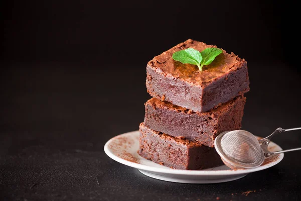 Chocolate brownie square pieces in stack on white plate with walnuts, decorated with mint leaves and cocoa on black background. Delicious dessert. Dark mood. Close up photography. Selective focus — Stock Photo, Image