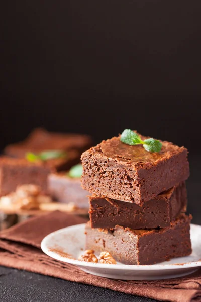Chocolate brownie square pieces in stack on white plate with walnuts, decorated with mint leaves and cocoa on black background. Delicious dessert. Dark mood. Close up photography. Selective focus — Stock Photo, Image
