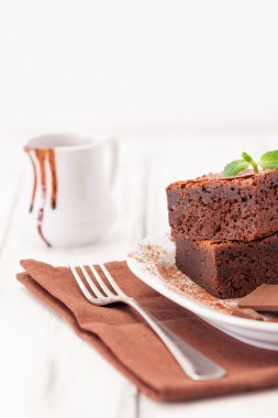 Çikolatalı kek kare parçalar halinde beyaz plaka üzerine nane yaprakları ve beyaz vintage ahşap zemin üzerine kakao tozu ile dekore edilmiştir. Amerikan geleneksel lezzetli tatlı. Fotoğraf kadar kapatın