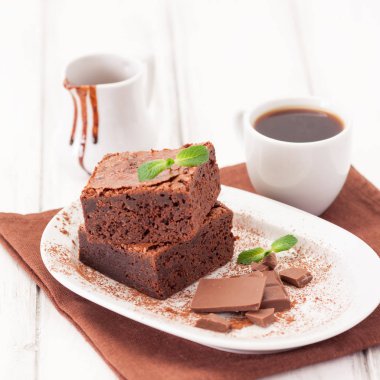 Çikolatalı kek kare parçalar halinde beyaz plaka üzerine nane yaprakları ve beyaz vintage ahşap zemin üzerine kakao tozu ile dekore edilmiştir. Amerikan geleneksel lezzetli tatlı. Fotoğraf kadar kapatın