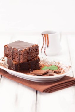 Çikolatalı kek kare parçalar halinde beyaz plaka üzerine nane yaprakları ve beyaz vintage ahşap zemin üzerine kakao tozu ile dekore edilmiştir. Amerikan geleneksel lezzetli tatlı. Fotoğraf kadar kapatın