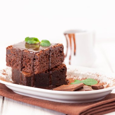 Çikolatalı kek kare parçalar halinde beyaz plaka üzerine nane yaprakları ve beyaz vintage ahşap zemin üzerine kakao tozu ile dekore edilmiştir. Amerikan geleneksel lezzetli tatlı. Fotoğraf kadar kapatın