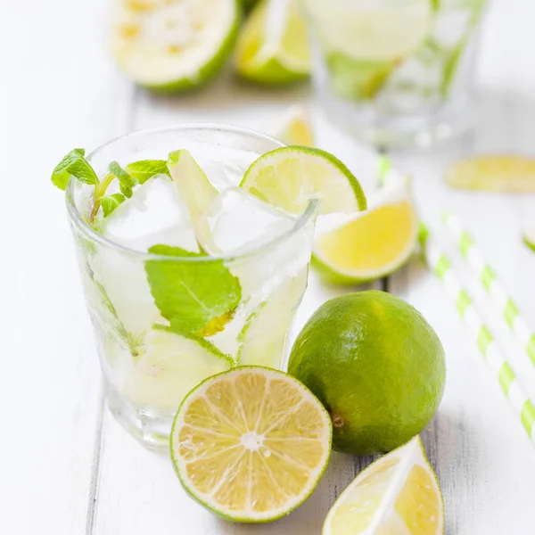 Caipirinha, mojito cocktail with lime, brown sugar, ice and mint leaves in beautiful glasses, cut green citrus on white wooden background. Summer alcohol drink. Close up photography. Selective focus — Stock Photo, Image