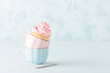 Pastel mavi zemin üzerine iki su bardağı hafif pembe krem süslemeli cupcake.