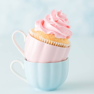 Pastel mavi zemin üzerine iki su bardağı hafif pembe krem süslemeli cupcake.