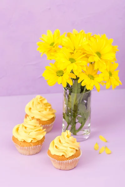 Crisântemo amarelo em vidro e cupcake com creme amarelo em fundo pastel violeta. Projeto de layout de dia da mãe com flores e sobremesa . — Fotografia de Stock