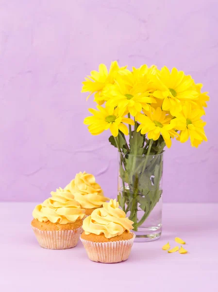 Crisântemo amarelo em vidro e cupcake com creme amarelo em fundo pastel violeta. Projeto de layout de dia da mãe com flores e sobremesa . — Fotografia de Stock