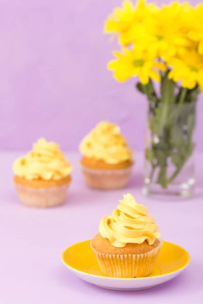 Κίτρινο χρυσάνθεμο σε γυαλί και cupcake με κίτρινη κρέμα σε βιολετί παστέλ φόντο. Ημέρα της μητέρας διάταξη σχεδιασμός με λουλούδια και επιδόρπιο. — Φωτογραφία Αρχείου