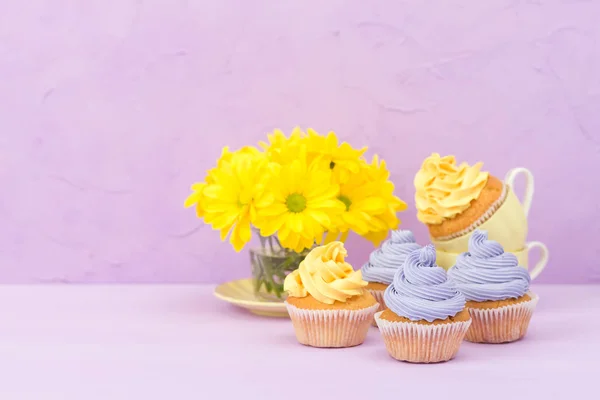 Cupcakes decorados com creme amarelo e violeta e crisântemos em fundo pastel violeta para cartão de saudação com copyscape . — Fotografia de Stock