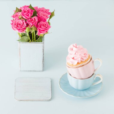 Pastel mavi zemin üzerine iki su bardağı hafif pembe krem süslemeli cupcake.