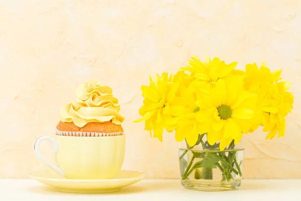 Кекс з ніжно-жовтим кремом та букетом з хризантем у склянці на жовтому пастельному фоні. Концепція натюрморту та мінімалізму — стокове фото