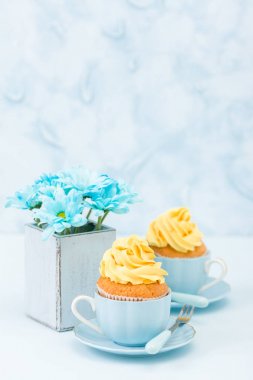 Cupcake ihale sarı krem dekorasyon ve retro eski püskü şık vazoda pastel mavi zemin üzerine mavi papatya buketi ile.