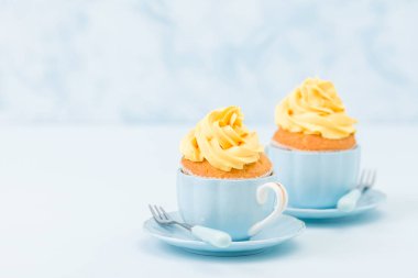Pastel mavi zemin üzerine iki küçük bardak tatlı sarı krem süslemeli cupcake.
