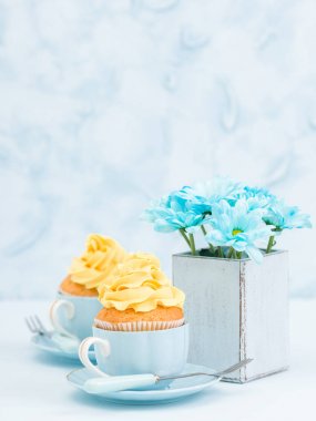 Cupcake ihale sarı krem dekorasyon ve retro eski püskü şık vazoda pastel mavi zemin üzerine mavi papatya buketi ile.