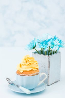 Cupcake ihale sarı krem dekorasyon ve retro eski püskü şık vazoda pastel mavi zemin üzerine mavi papatya buketi ile.