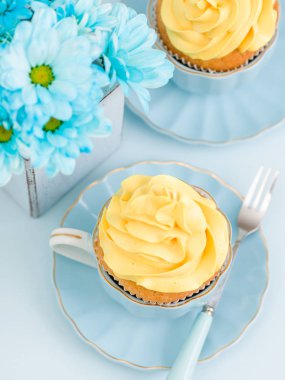 Cupcake ihale sarı krem dekorasyon ve retro eski püskü şık vazoda pastel mavi zemin üzerine mavi papatya buketi ile.