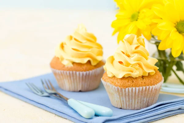 Cupcake with yellow cream decoration on blue napkin and bouquet of yellow chrysanthemum in small glass.