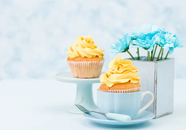 Cupcakes with tender yellow cream decoration and bouquet of blue chrysanthemum in retro shabby chic vase on blue pastel background.