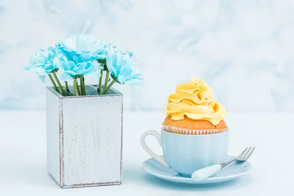 Cupcake with tender yellow cream decoration and bouquet of blue chrysanthemum in retro shabby chic vase on blue pastel background. — Stock Photo, Image