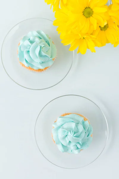 Cupcake with sweet blue cream decoration on sauser and bouquet of yellow chrysanthemum in glasses cup. Flat-lay concept