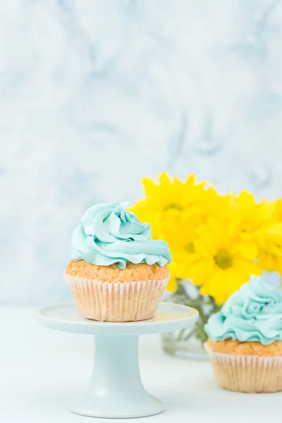 Cupcake with blue cream decoration on vintage stand and bouquet of yellow chrysanthemum in glasses vase.