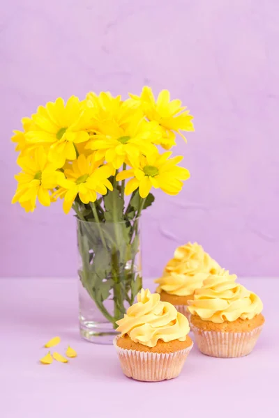 Crisântemo amarelo em vidro e cupcakes doces com decoração em fundo pastel violeta . — Fotografia de Stock