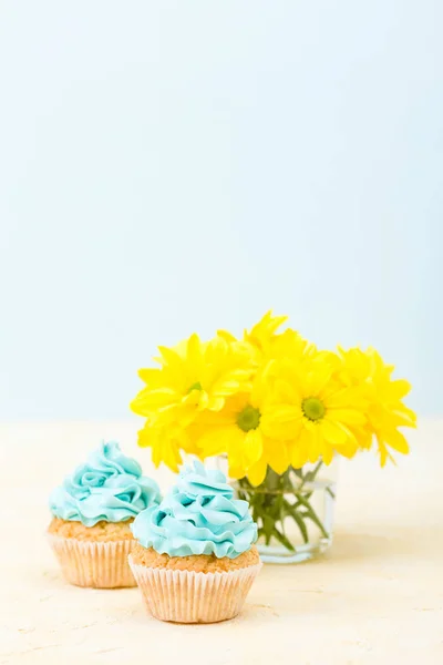 Mavi krem dekorasyon ve cam pastel zemin üzerine sarı Kasımpatı buket ile cupcakes. — Stok fotoğraf