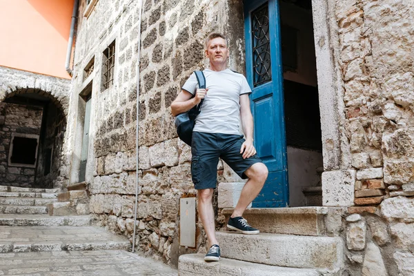 Stylish tourist. Man dressed in a white shirt and shorts with backpack over his shoulder. Standing on the steps of European city