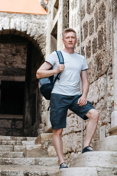 Stylish tourist. Man dressed in a white shirt and shorts with backpack over his shoulder. Standing on the steps of European city