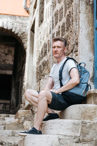 Stylish tourist. Man dressed in a white shirt and shorts with backpack over his shoulder. Sitting on the steps of European city