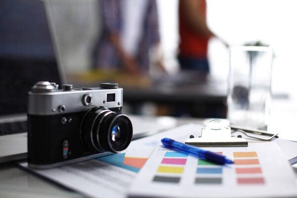 Ноутбук и камера на столе, два бизнесмена стоят на заднем плане
