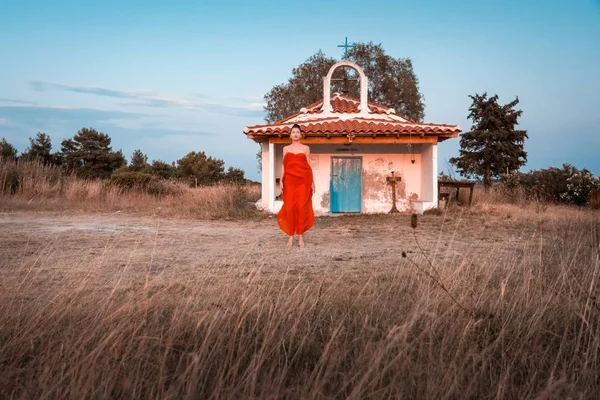 Belo modelo jovem em um vestido vermelho em Pefkochori, Kassandra, Chalkidiki, Grécia com uma pequena igreja ortodoxa em um contexto no verão - captura artística — Fotografia de Stock