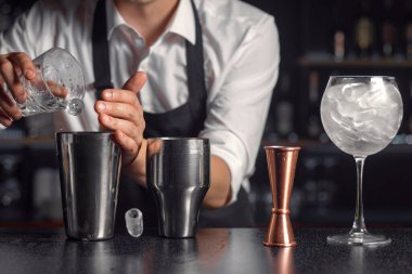 Soğuk içecek hazırlığı aşaması. Barmenin elleri içecek karıştırıcısına buz ekliyor. Yüzeyde buz küpü var. Karıştırıcının yanında duran buzla doldurulmuş ölçü bardağı ve şarap kadehi. Kapat.