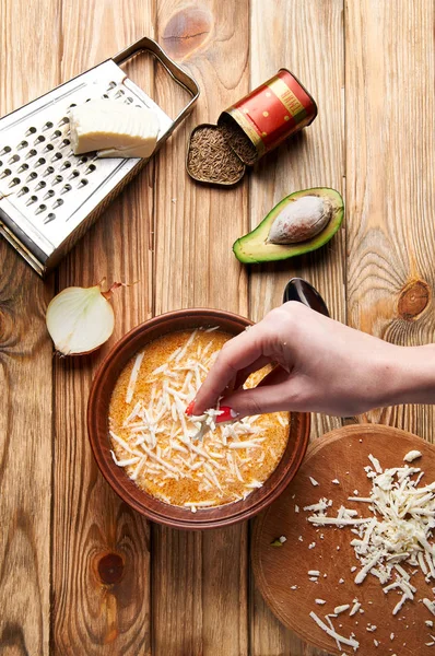 La mano femenina espolvorea con queso deliciosa sopa de crema argentina. En la vieja mesa de madera se encuentra un gran rallador, queso, un plato de sopa, cebollas en rodajas, aguacate y comino . — Foto de Stock