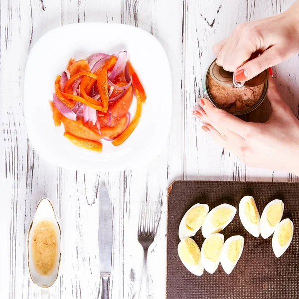 Cocinar preparando ensalada. La mujer abre un frasco de carne enlatada en la cocina. La chica va a poner el atún en ensalada con tomates, cebollas y pimientos. Ensalada aderezo y huevos cocidos en el fondo . — Foto de Stock