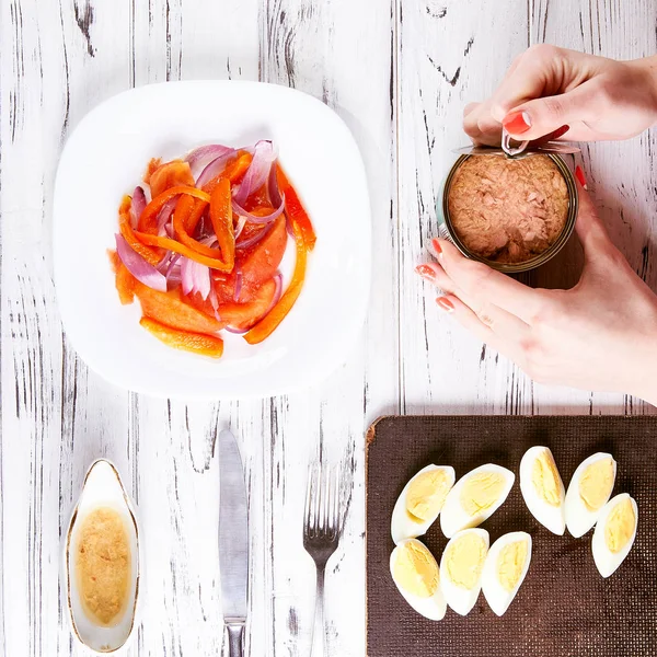 Koch bereitet Salat zu. Frau öffnet in der Küche ein Glas mit Fleischkonserven. Das Mädchen legt den Thunfisch in Salat mit Tomaten, Zwiebeln und Paprika. Salatdressing und gekochte Eier im Hintergrund. — Stockfoto