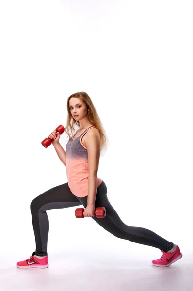 Young beautiful sporty woman with a dumbbell doing lunges. Slim girl is engaged in fitness. Fitness trainer in leggings, a sports T-shirt, sneakers with red dumbbells. Fitness training. — Stock Photo, Image