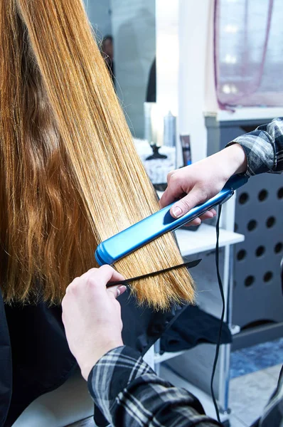 Professional hairdresser straightens model hair. Hair iron for hair straightening. Woman with beautiful long thick blond hair. Girl in a barber\'s chair at the beauty salon.