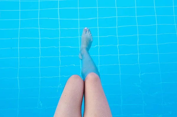 The girl splashes her legs in the pool with clear blue water. A woman is resting by the pool on a hot summer day. Shapely female legs. Women's legs and feet in water.