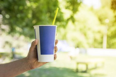 Elini kağıt bardak su, kahve veya çay genel parkta tutuyor, ağaç bir arka plan ve bokeh