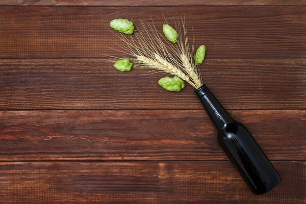 Beer in the form of hops and ears of wheat pours from the neck of the bottle. A bottle of beer with green hops and wheat spikelets on wooden background. — Stock Photo, Image