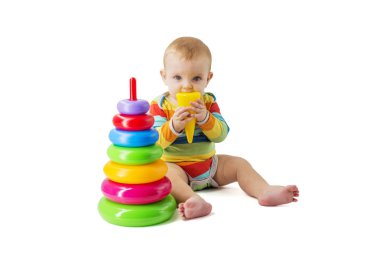 Tulum giymiş sevimli bir bebek beyaz arka planda oturan renkli plastik bir piramitle oynuyor. Bir çocuk renkli bir piramit toplar