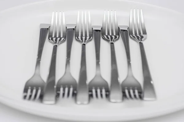 Muitos garfos inoxidáveis brilhantes estão em uma placa branca na mesa. .. Louça de cerâmica e talheres simples — Fotografia de Stock