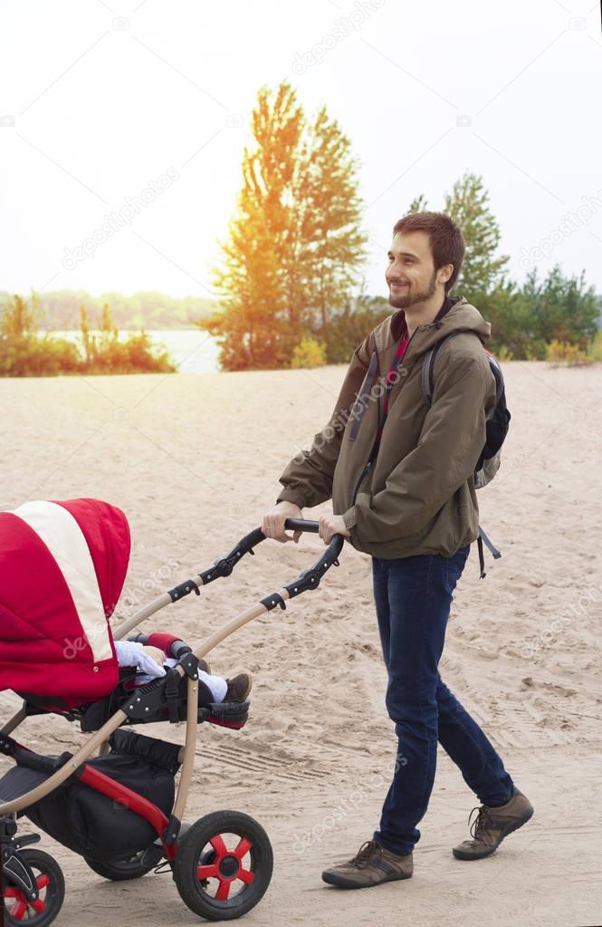 Young father helps mother, he walks with a baby carriage in the park by the river