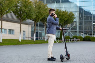 Günbatımında havaalanının yanındaki kaldırımda duran ve akıllı telefondan konuşan, e-scooterlı genç yakışıklı bir adam. Modern elektrikli scooterda trend şehir ulaşımı. Çevre dostu hareketlilik kavramı.