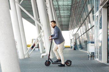 Genç yakışıklı adam e-scooter 'la havaalanından ayrılıyor. Yolculukta bir nakliye aracı kirala. Modern elektrikli scooter 'la son moda şehir ulaşımı. Çevre dostu hareketlilik kavramı.