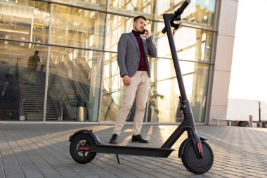 Günbatımında havaalanının yanındaki kaldırımda duran ve akıllı telefondan konuşan, e-scooterlı genç yakışıklı bir adam. Modern elektrikli scooterda trend şehir ulaşımı. Çevre dostu hareketlilik kavramı.